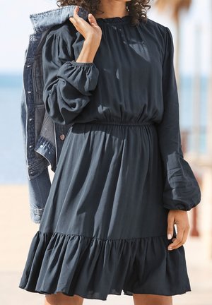 Frau in einem langärmligen dunklen Kleid, die eine Jeansjacke über der Schulter hält und im Freien in der Nähe eines Strandes steht.