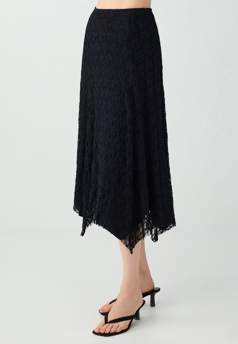 Jupe midi en dentelle noire avec un ourlet asymétrique portée avec des sandales à talons noires sur un fond clair.