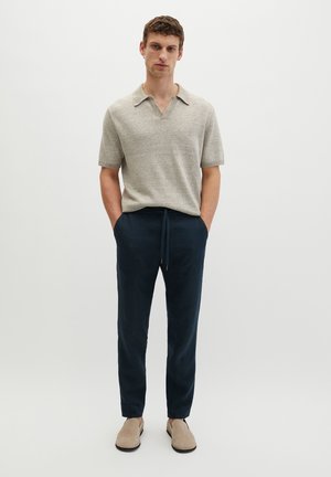 Jonge man staat met zijn handen in zijn zakken, draagt een lichtgrijs kort mouw gebreid polo, donkere marineblauwe broek met trekkoord en beige schoenen.