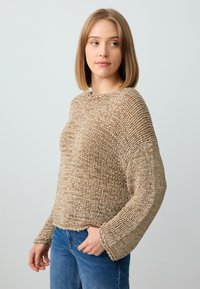 Beige gestrickter Pullover mit weiten Ärmeln und lockerer Passform. Strukturiertes Material mit Rippdesign, getragen über blauen Jeans.