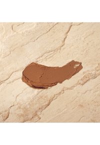 Brown makeup cream smeared on a light beige, textured surface with subtle veining. The cream has a smooth, thick consistency.
