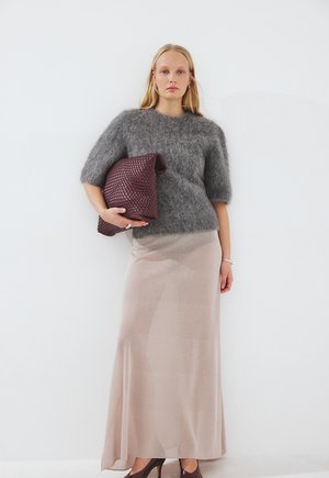 Mujer con un suéter corto de manga corta gris peludo, falda larga beige, tacones marrones, sosteniendo un gran bolso de mano tejido color burdeos sobre un fondo blanco.