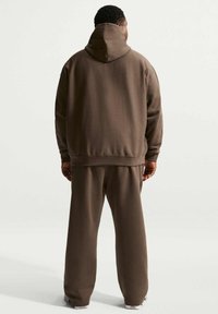 Sweat à capuche en coton marron avec capuche ajustable, poignets et taille côtelés, assorti à un pantalon à coupe décontractée. Texture lisse, design minimaliste.