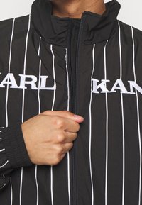 Svart jacka med vita vertikala ränder, hög krage, dragkedja och broderat varumärke på framsidan; textil med strukturerad yta och elastiska muddar.