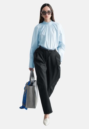Femme portant une blouse bleu clair oversize, un pantalon noir taille haute, des chaussures blanches, de grandes lunettes de soleil, portant un sac fourre-tout gris avec une écharpe bleue.