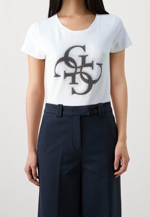 Vrouw draagt een wit T-shirt met een zwart in elkaar grijpende cirkelpatroon en donkerblauwe broek met hoge taille.