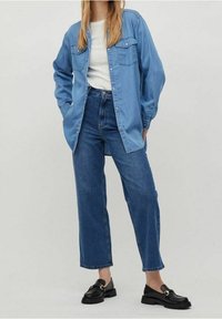 Chemise en jean bleu clair avec deux poches poitrine, associée à un jean bleu foncé taille haute et des mocassins noirs. Texture lisse, coupe décontractée.