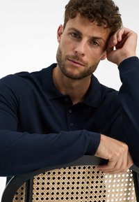 Polo a maniche lunghe blu navy con tessuto textured, caratterizzato da colletto e bottoni, indossato da una persona che si riposa sullo schienale di una sedia in rattan.
