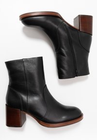 Botines de cuero negro con tacón grueso de madera y cremallera lateral, que presentan una textura suave y un diseño aerodinámico.