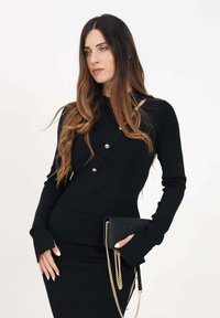 Vestido negro de punto con silueta ajustada, que presenta botones dorados y mangas largas. Acompañado de un pequeño bolso negro de mano con cadena.