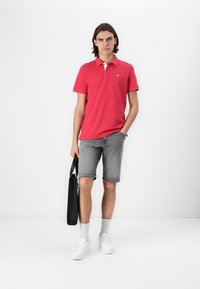 T-shirt polo rouge avec des accents blancs, associé à un short en denim gris. Baskets blanches et chaussettes grises, tenant un sac noir. Tenue simple et moderne.