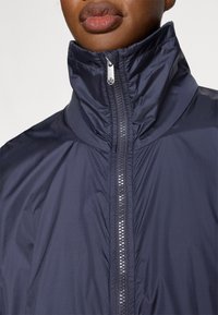 Granatowa kurtka z wysokim kołnierzem, wyposażona w zamek błyskawiczny na całej długości oraz teksturowaną tkaninę. Minimalistyczny design z odblaskowymi elementami sprzętu.