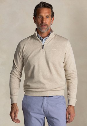 Beige kwart-rits pullover met ribbelmanchetten en -zoom, zachte textuur, met een ritssluiting en subtiel logo op de borst, gecombineerd met lichtblauwe broek.