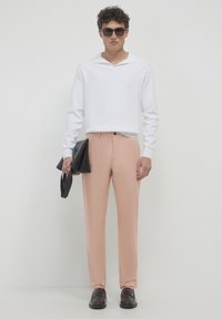 Helle rosa Hose mit schmaler Passform, kombiniert mit einem weißen Langarm-Polo, schwarzen Sonnenbrillen und einer schwarzen Handtasche, vor einem schlichten Hintergrund.