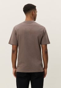 Καφέ βαμβακερό μπλουζάκι με κοντά μανίκια και στρογγυλή λαιμόκοψη, με απλό σχέδιο και λεία υφή. Η πίσω όψη δεν έχει γραφικά ή σχέδια.