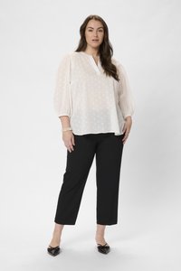 Blouse blanche à pois noirs avec un décolleté en V et des manches bouffantes, associée à un pantalon noir raccourci et des chaussures noires plates. Tissu texturé.