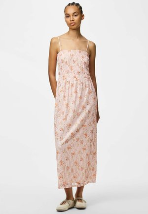 Femme portant une robe longue sans manches à fleurs roses avec des bretelles spaghetti et des chaussures plates beiges, debout devant un fond blanc uni.