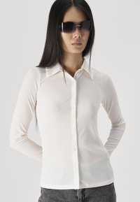 Long-sleeved, white ribbed shirt with a button-down front and collar. Model wearing dark sunglasses; textured fabric visible.