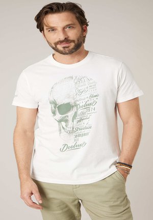Homme barbu portant un t-shirt blanc avec un motif de crâne vert et un texte mixte, pantalon beige et bracelets de perles au poignet droit.