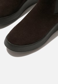 Botas de tobillo de ante marrón con punta redondeada, suela de goma negra y textura suave. Presentan un diseño minimalista y sin acentos visibles.