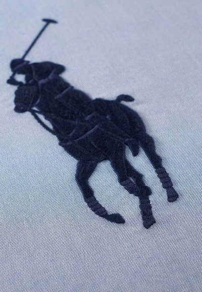 Μπλε ναυτικό κέντημα ενός παίκτη πόλο και αλόγου σε ανοιχτό μπλε ύφασμα, που παρουσιάζει λεπτομερείς ραφές και υφή επιφάνειας.