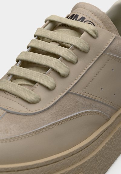 Μπεζ αθλητικό παπούτσι με χοντρή σόλα, πάνελ από σουέτ και δέρμα, φαρδιά επίπεδα κορδόνια και ραμμένες λεπτομέρειες.