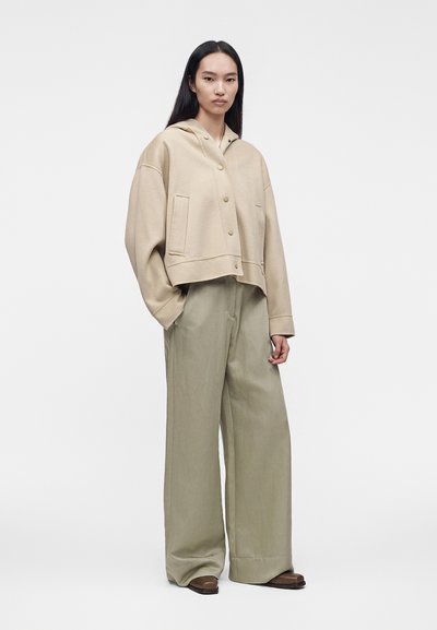 Personne aux longs cheveux foncés porte une veste beige boutonnée avec des manches larges et un pantalon ample vert olive clair sur un fond clair uni.