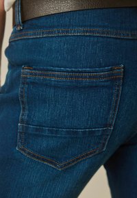 Jeans de mezclilla azul oscuro con un bolsillo trasero que presenta costuras beige y un acabado texturizado. El diseño enfatiza una forma ajustada.