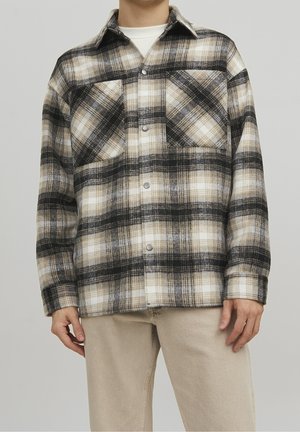 Person trägt beige Hose und eine beige, schwarz-weiß karierte, geknöpfte Jacke über einem weißen Hemd vor einem einfarbigen Hintergrund.