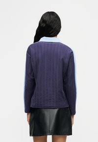 Γυναίκα με μακριά μαύρα μαλλιά φοράει ένα μπλε σκούρο πλεκτό πουλόβερ με γαλάζια μανίκια και γιακά, συνδυασμένο με μαύρη δερμάτινη φούστα, γυρισμένη προς τα πίσω.