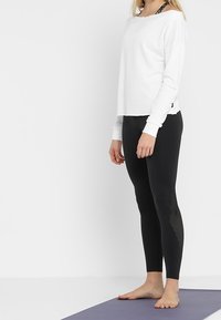 Bílé tričko s dlouhým rukávem a širokým výstřihem, spárované s černými legínami se síťovinovými detaily na vnější straně. Oba kousky jsou těsně přiléhavé.