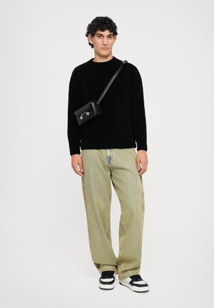 Man wearing black sweater, wide light green jeans, black-and-white sneakers, and a small black crossbody bag against white background.