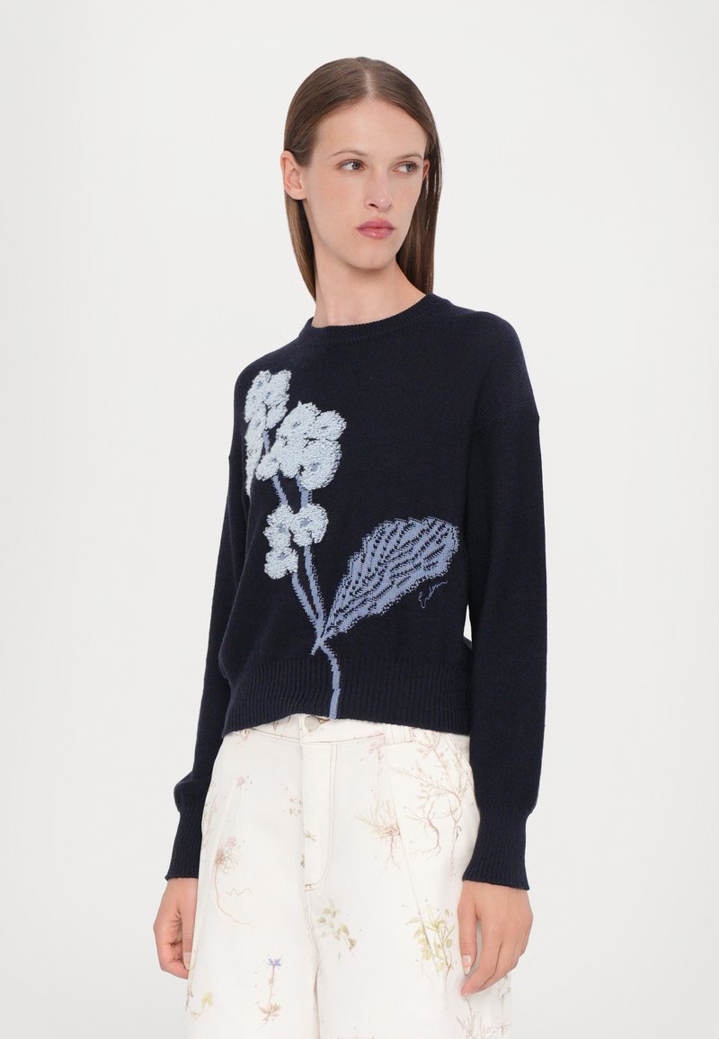 Pull en maille marine à manches longues, avec une broderie fleurie bleu clair et un ourlet côtelé. Associé à un pantalon crème à motif floral.