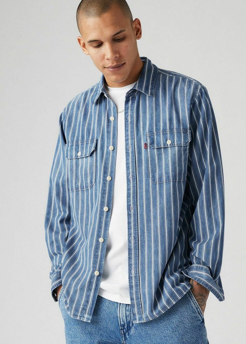 Chemise en denim bleu avec des rayures verticales blanches, deux poches poitrine et fermeture à boutons, portée sur un t-shirt blanc, associée à un jean bleu.