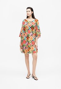 Γυναίκα που στέκεται φορώντας ένα φόρεμα μέχρι το γόνατο με πολύχρωμο λουλουδένιο σχέδιο και φτερωτά μανίκια, σε συνδυασμό με μαύρα πέδιλα με λουράκια.