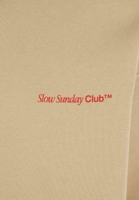 Beige bomuldsstof med ribbet tekstur har en rød broderet "Slow Sunday Club™" logo centreret på brystet.