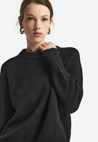 Mörk stickad tröja med ribbad krage och ärmslut, med en lös passform och strukturerad mönstring. Tyget ser varmt och mysigt ut.