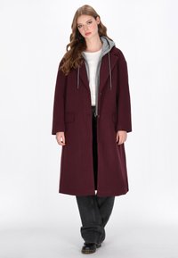 Cappotto lungo bordeaux con una fodera grigia con cappuccio, dotato di chiusura a bottoni, due tasche frontali e una vestibilità rilassata, abbinato a pantaloni neri.