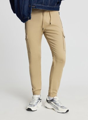 Beżowe spodnie joggery typu cargo z bocznymi kieszeniami, elastycznymi mankietami, ściągaczem w pasie, zestawione z białymi i szarymi sportowymi sneakersami.