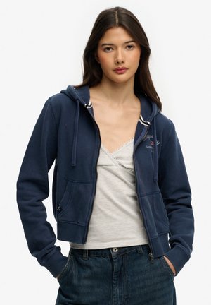 Young woman with long dark hair wearing navy zip-up hoodie over light gray camisole and dark jeans, hands in pockets, neutral expression.