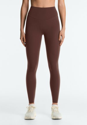 Femme portant des leggings marron taille haute et des baskets blanches, debout avec les bras détendus le long du corps devant un fond uni.