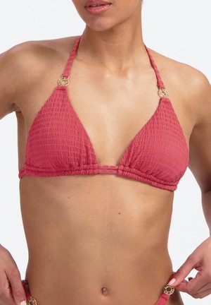 Frau trägt einen strukturierten roten Triangel-Bikini-Top mit goldenen Ringakzenten und justiert den passenden Bikinihöschenriemen an ihrer Hüfte.