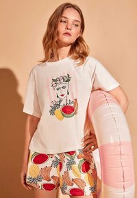 Jeune femme portant un t-shirt blanc avec un imprimé visage et fruits, un short assorti à motifs fruits, tenant une bouée rose et blanche, debout devant un fond beige.