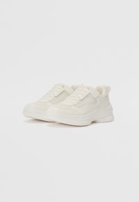 Baskets blanches avec une tige en maille texturée et en cuir, fermeture à lacets et une semelle épaisse. Présente un subtil accent doré près des lacets.