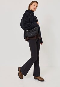 Black nylon bag with silver zipper, worn crossbody; model in a black oversized hoodie, dark flared pants, and leopard print ankle boots.