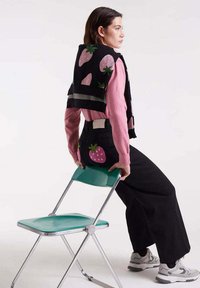 Mujer vestida con un suéter rosa y pantalones negros con estampados de fresas se apoya en una silla plegable verde contra un fondo blanco.