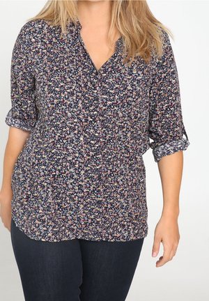 Mujer que lleva una camisa abotonada de manga larga con estampado floral oscuro, con las mangas remangadas hasta los codos y pantalones azul oscuro.