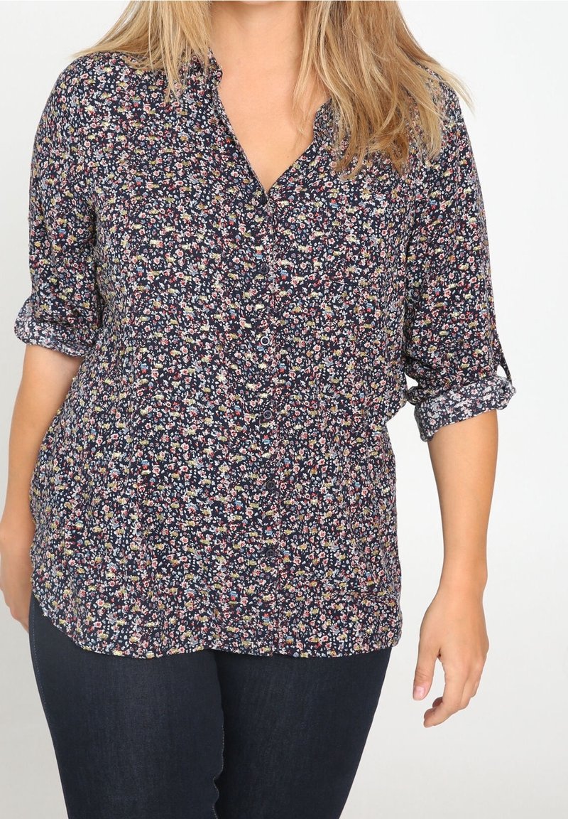 Femme portant une chemise boutonnée à manches longues, à motif floral foncé, manches retroussées jusqu’aux coudes, et un pantalon bleu foncé.