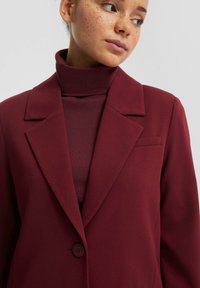 Blazer borgogna con colletto a scesa, chiusura a singolo bottone e una piccola tasca sul petto. Tessuto morbido, abbinato a un maglione a collo alto coordinato.