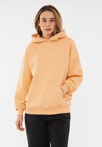 Felpa con cappuccio color pesca, dotata di tasca a marsupio, polsini a costine e orlo ondulato. Realizzata in tessuto morbido con una vestibilità comoda.